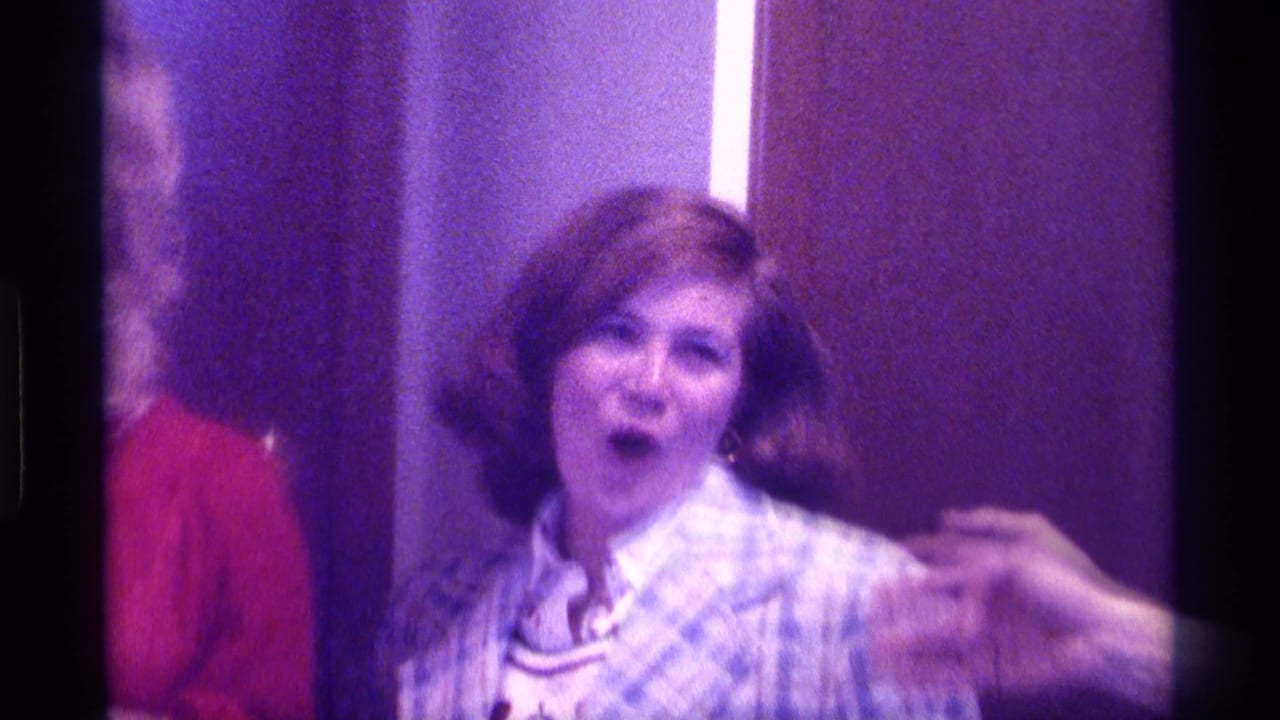 Woman in a plaid blazer reacts with her mouth open in a purple-lit interior.