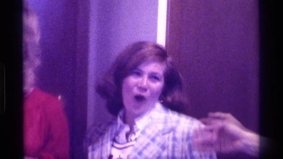 In 1976 Kentucky home-movie footage, a woman in a plaid blazer reacts while a hand reaches near her shoulder indoors.