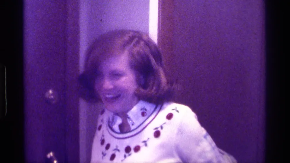 A woman in a white sweater laughs while leaning forward, with the plaid blazer sliding off in the 1976 Kentucky home movie.