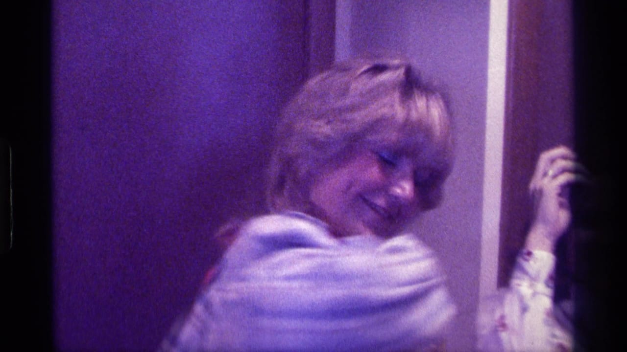 In a 1976 Kentucky home-movie frame, a blonde woman in a light plaid jacket smiles near an interior doorway as another person stands nearby.