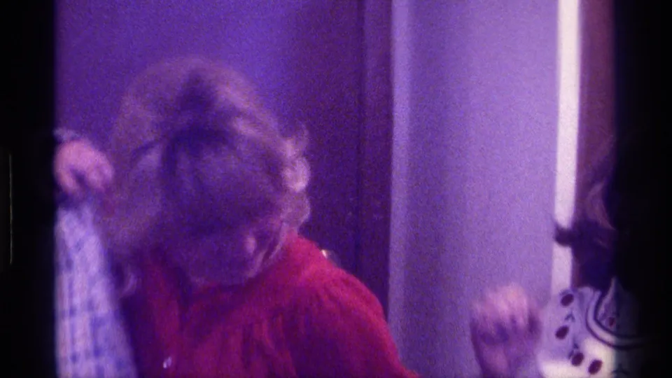 Two women appear in a close indoor 1976 Kentucky home-movie frame, with one in a red shirt and plaid jacket looking downward as they laugh.