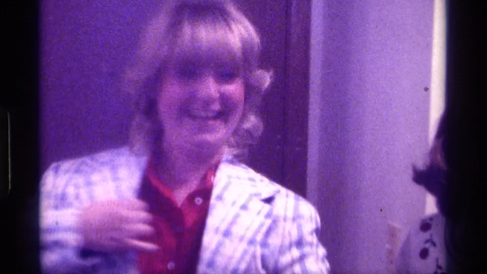 A woman in a red shirt and light plaid jacket smiles toward the camera in a grainy 1976 Kentucky indoor home-movie frame.