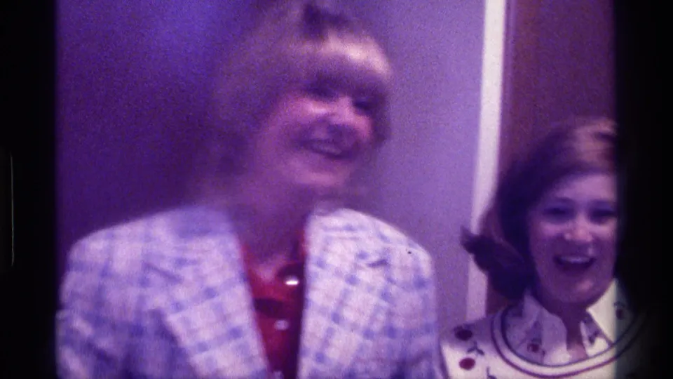 Two women laugh together indoors in a 1976 Kentucky home-movie frame, with visible 1970s clothing and soft purple 8mm color.