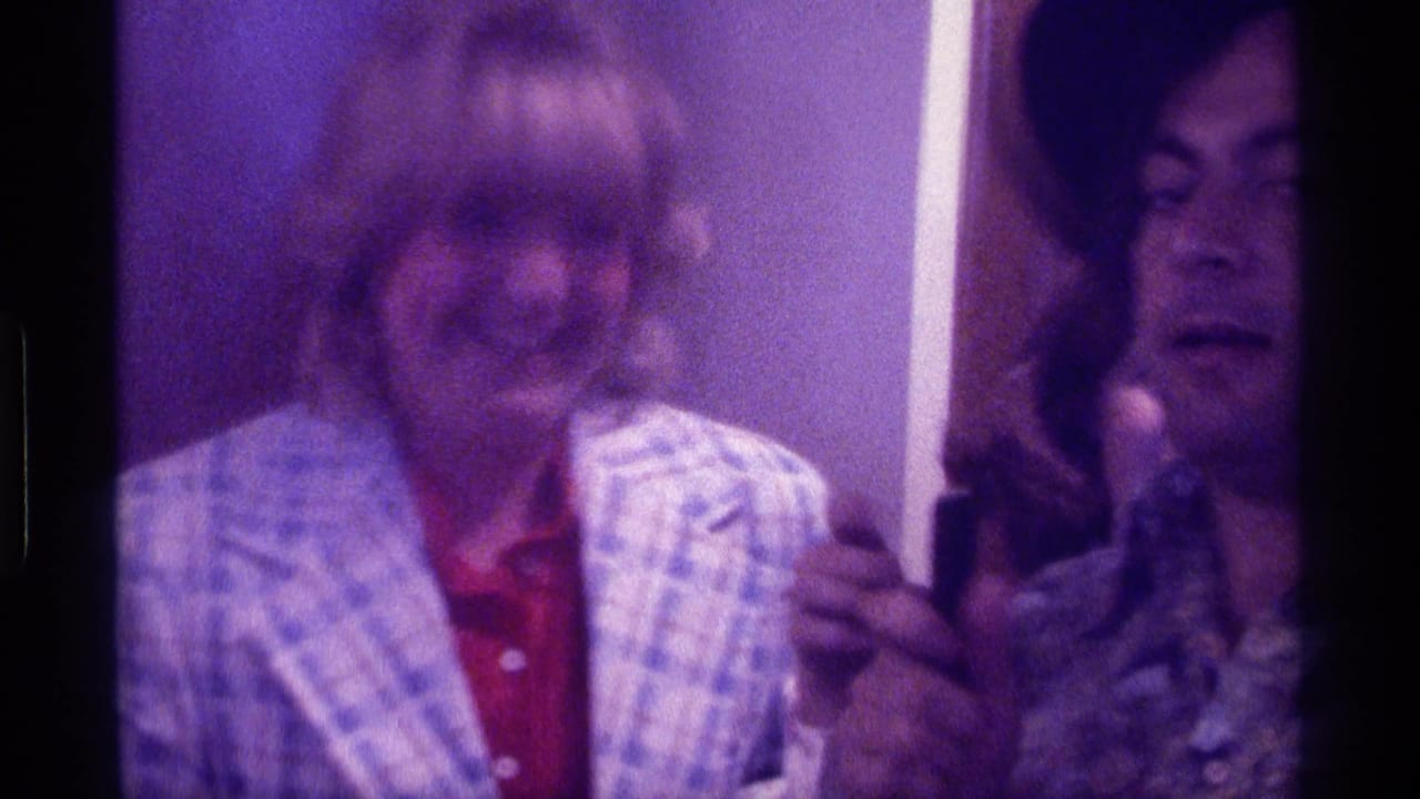 1976 Kentucky 8mm frame of a woman in a blue plaid jacket near a man in a black hat with a handheld microphone indoors.