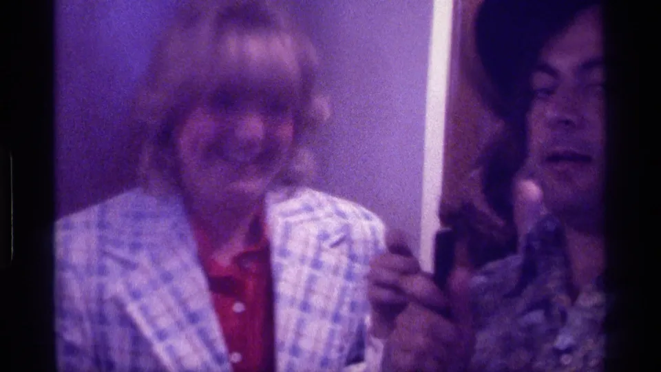 1976 Kentucky 8mm frame of a woman in a blue plaid jacket near a man in a black hat with a handheld microphone indoors.