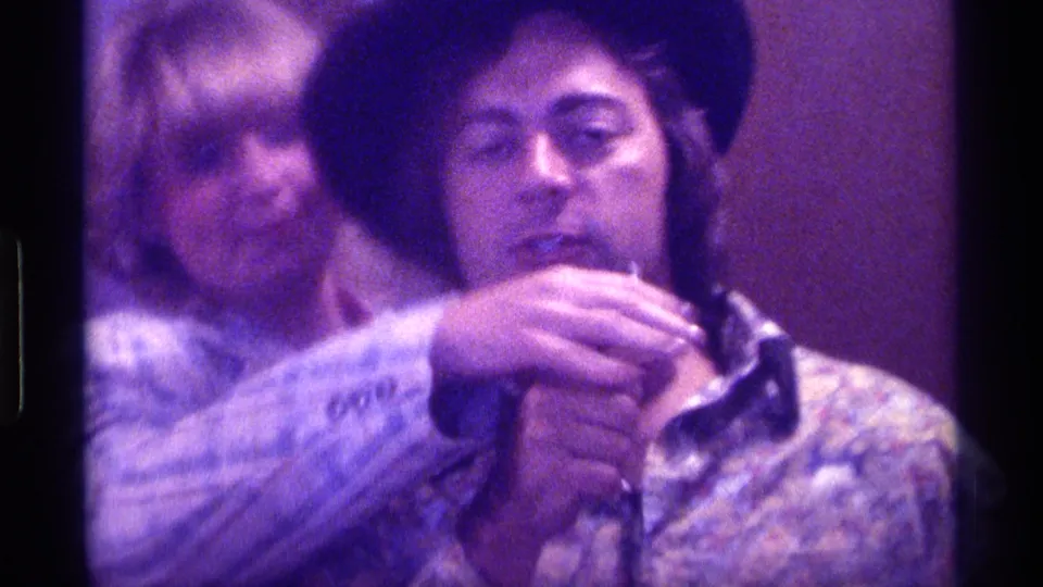 1976 Kentucky 8mm frame showing a man in a black hat and patterned shirt holding a microphone, with a woman behind him.