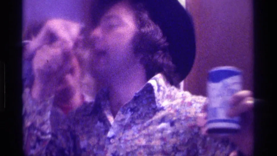 1976 Kentucky 8mm frame of a man in a black hat lifting his head with a cup in hand while a woman remains blurred behind him indoors.