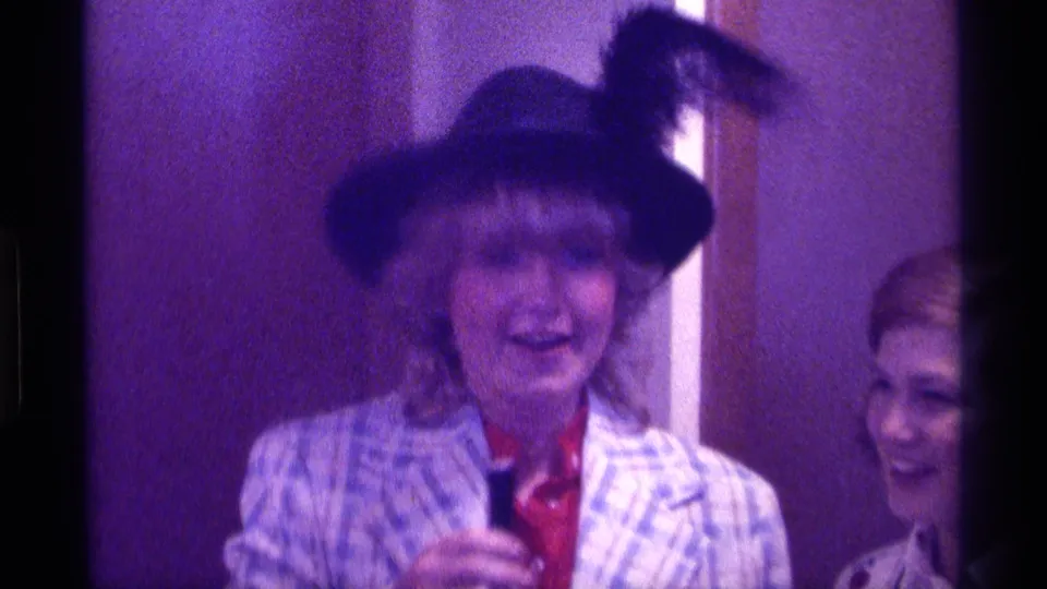 1976 Kentucky home-movie frame of a woman in a feathered hat and plaid blazer smiling in a purple-lit room.