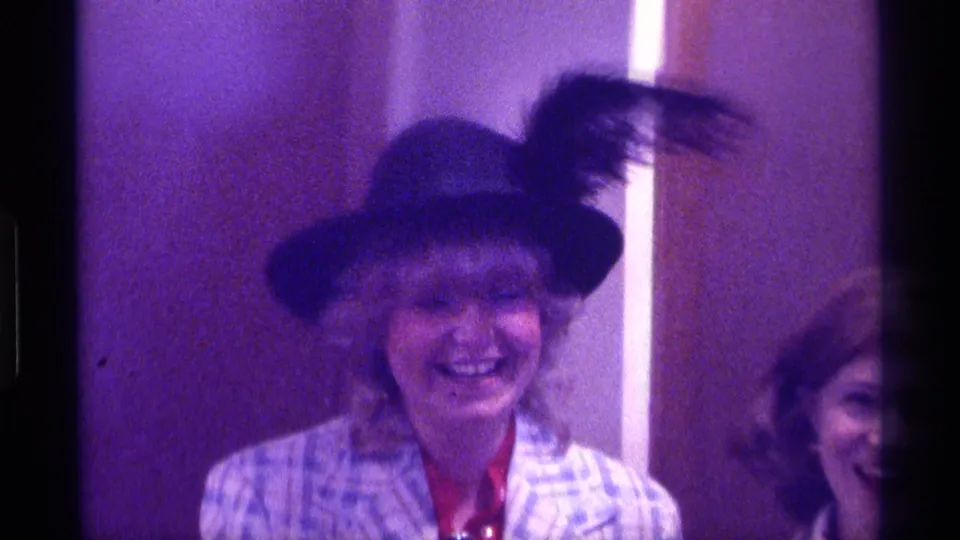 1976 Kentucky frame showing a woman in a feathered hat laughing in a soft, grainy 8mm interior shot.