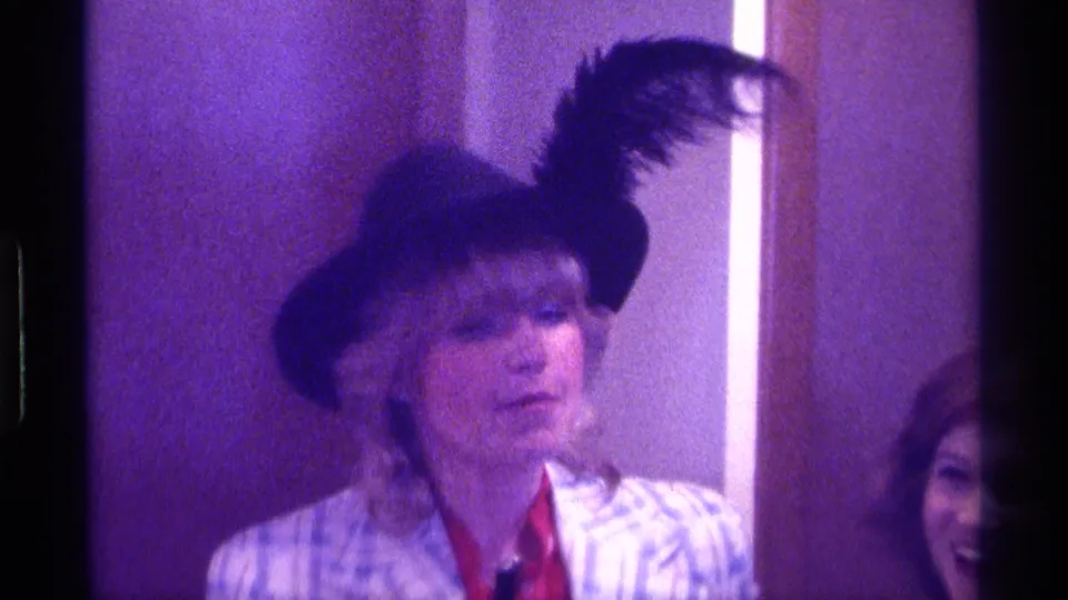 1976 Kentucky 8mm frame of a woman in a feathered hat looking aside in a dim private interior.