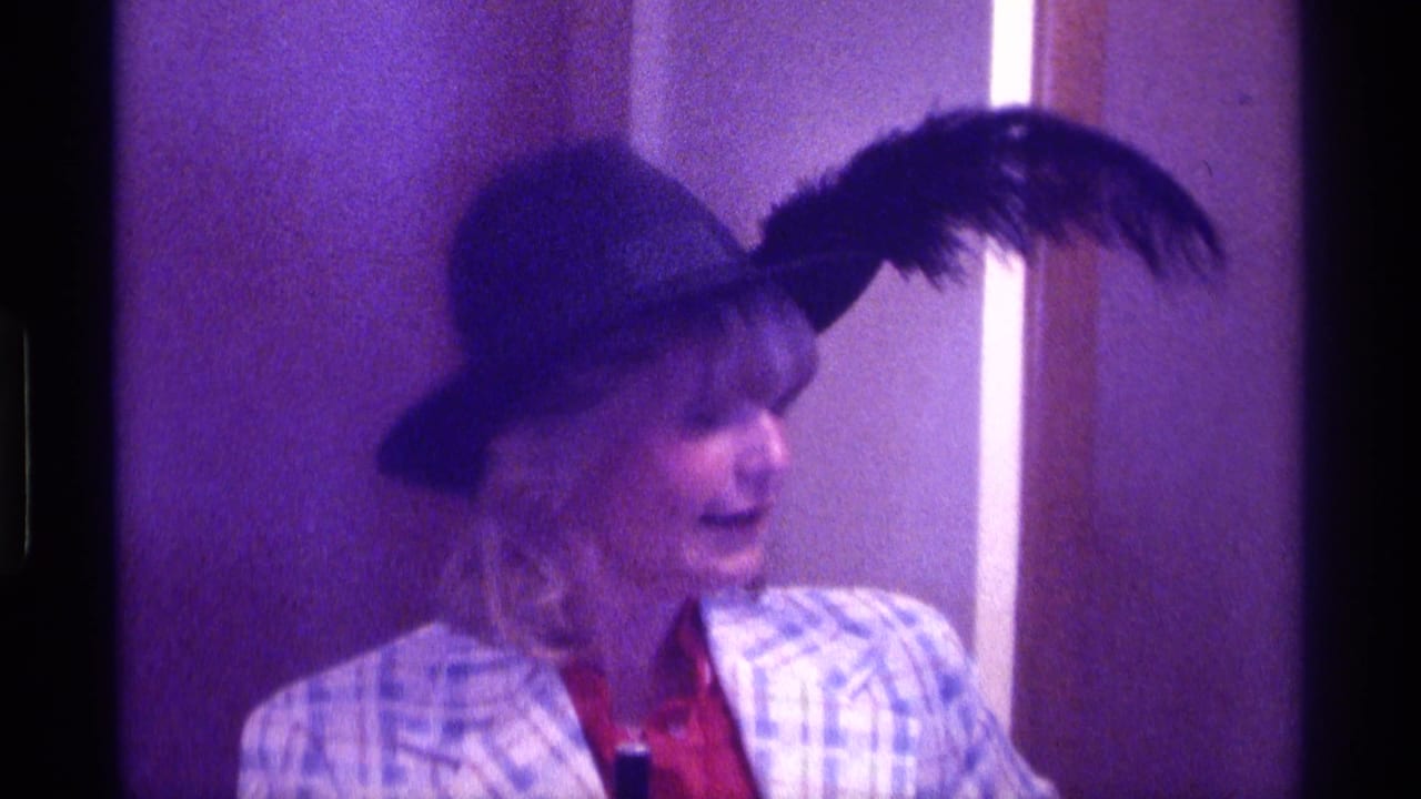 In this 1976 Kentucky 8mm frame, an adult woman in a feathered hat and plaid jacket looks off to the side indoors.