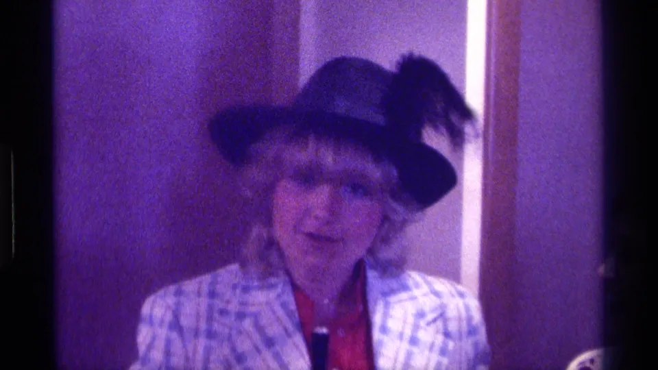 A 1976 Kentucky home-movie frame shows an adult woman facing the camera in a dark hat with a feather and a light plaid jacket.