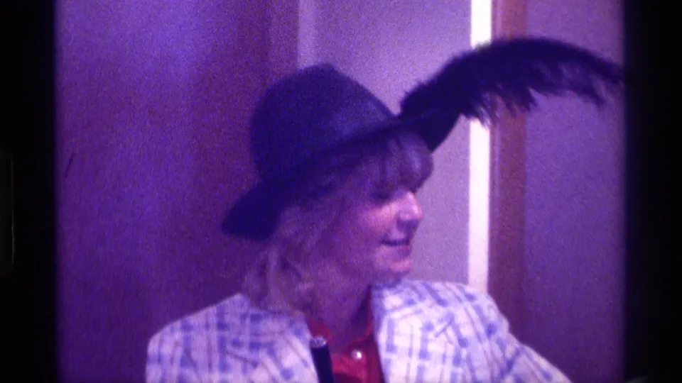 The woman smiles toward someone off camera in this 1976 Kentucky 8mm frame, with her feathered hat and plaid jacket prominent.
