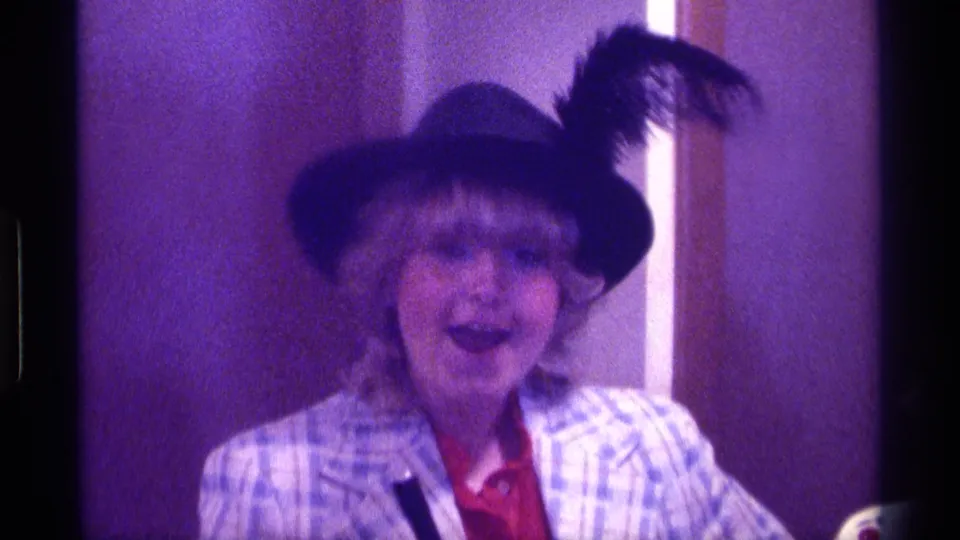 A 1976 Kentucky frame shows the woman facing the camera with an animated expression, wearing a feathered hat and plaid jacket.