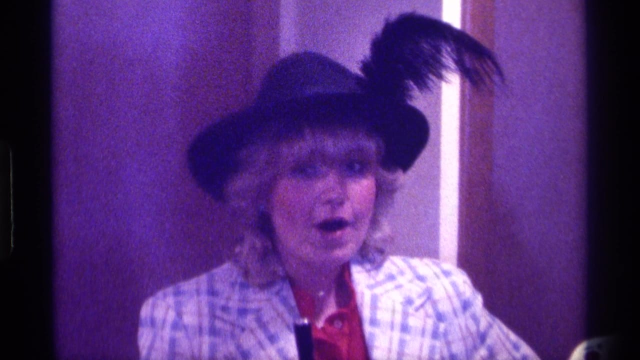 Woman in a feathered hat and plaid jacket faces the camera indoors.