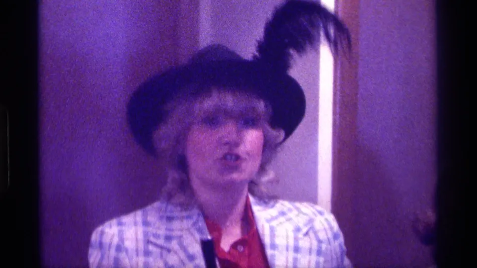The woman continues singing or speaking toward the camera, her feathered hat and plaid jacket visible in purple-tinted 8mm footage.