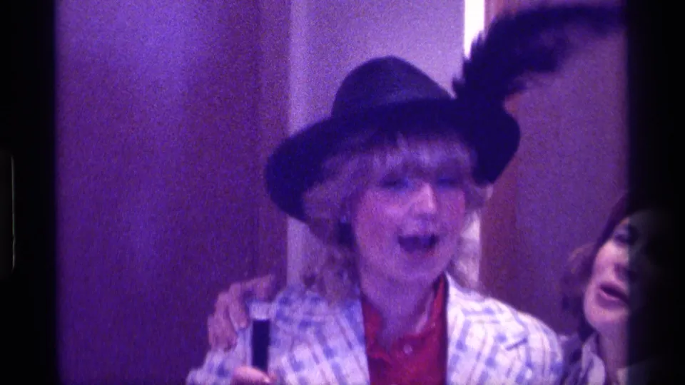 The singer appears joyful as another person enters beside her, both partially framed in a purple-tinted 1976 Kentucky 8mm party scene.
