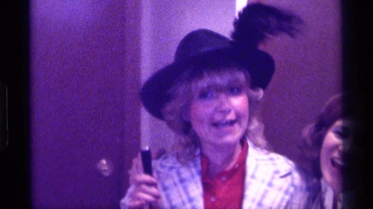 In Kentucky 1976 home-movie footage, a woman in a feathered hat and plaid jacket holds a microphone as another adult smiles behind her.