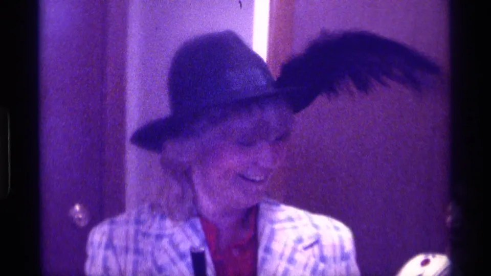 A woman in a feathered hat and plaid jacket turns to the side with a microphone in dim purple-tinted 1970s indoor footage.