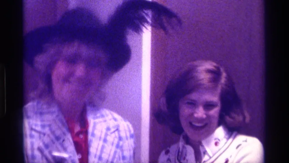 The woman in the feathered hat moves past a smiling woman in a light jacket during the Kentucky 1976 party footage.