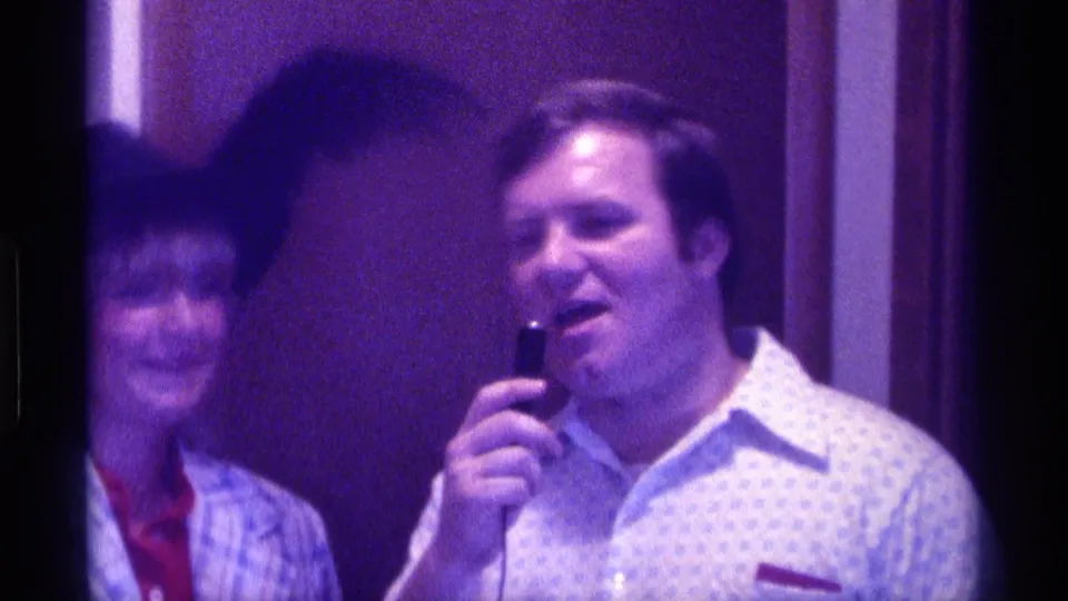 A man in a patterned shirt holds the microphone as the woman in the feathered hat smiles nearby in Kentucky 1976 home-movie footage.