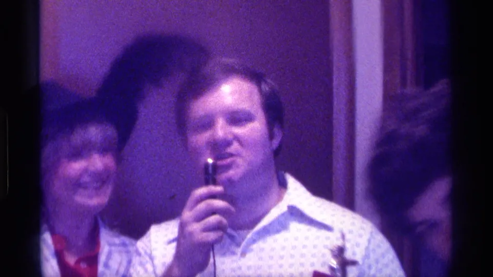 1976 Kentucky indoor gathering with a man using a microphone, a smiling woman in a feathered hat, and partial faces nearby.