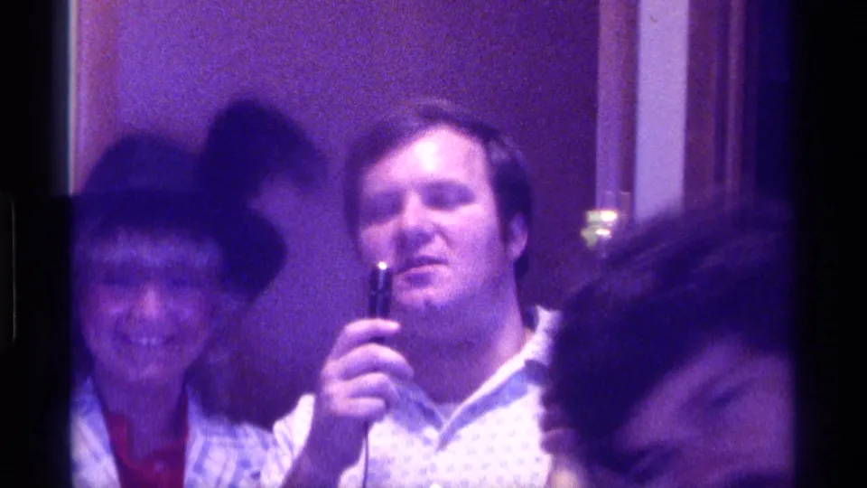 1976 Kentucky home-movie still with a smiling woman in plaid, a man with a microphone, and a foreground attendee partly blocking the view.