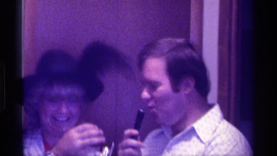 1976 Kentucky frame showing the man angled toward the smiling woman in a feathered hat during an indoor social exchange.