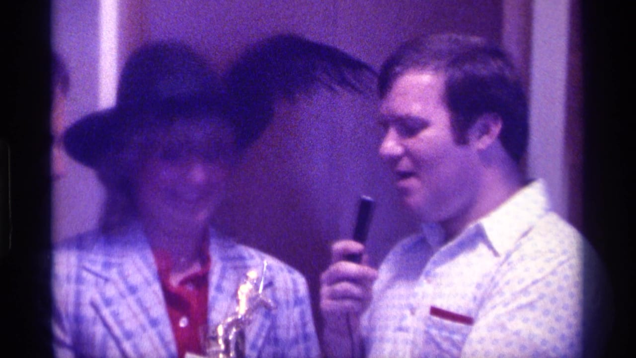 In 1976 Kentucky home-movie footage, a woman in a black hat and plaid jacket stands beside a man holding a microphone during an indoor celebration.