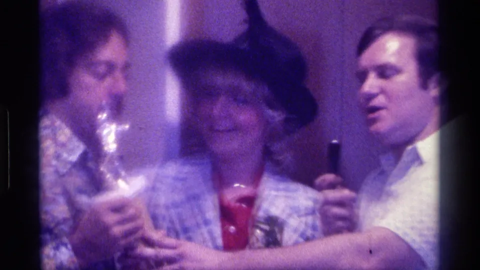 Adults in patterned shirts gather around a woman in a black feathered hat while a man holds a microphone in a close indoor 1970s scene.