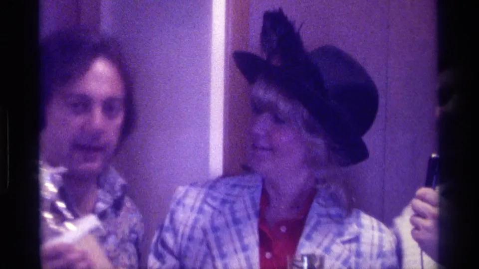 A man in a floral shirt stands close to the woman in a black hat and plaid jacket, with a microphone partly visible at the edge.