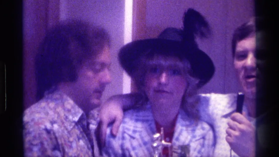 In the indoor 1976 Kentucky scene, a man leans toward the woman in a black hat as another man smiles and holds a microphone.