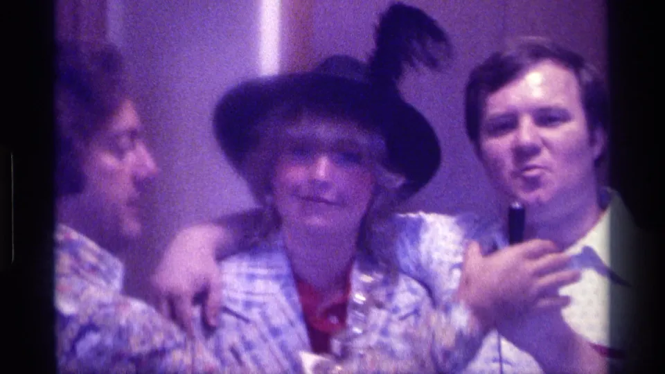 1976 Kentucky 8mm frame of adults standing close together indoors, with a woman in a black hat between men and a handheld microphone visible.