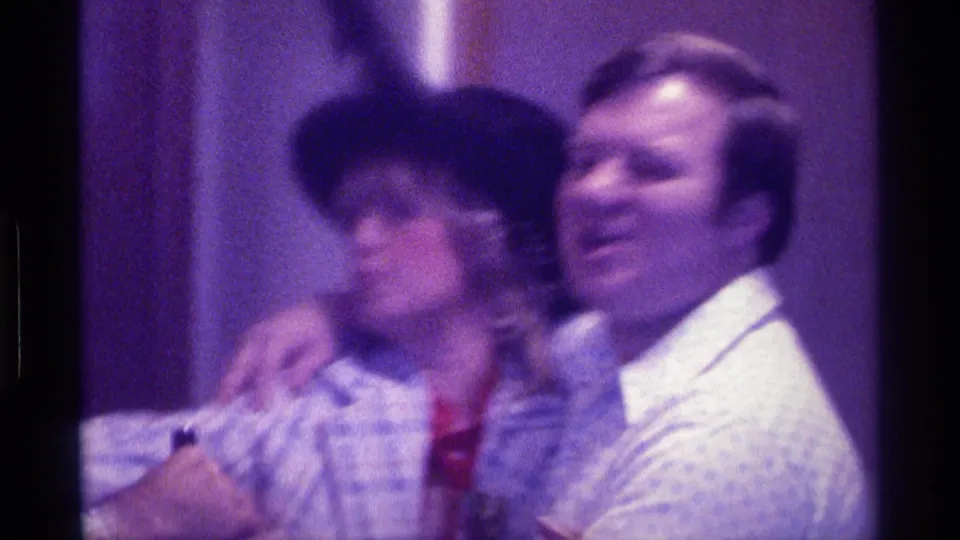 1976 Kentucky home-movie frame showing a woman in a black hat beside a smiling man, with arms around shoulders in an indoor room.