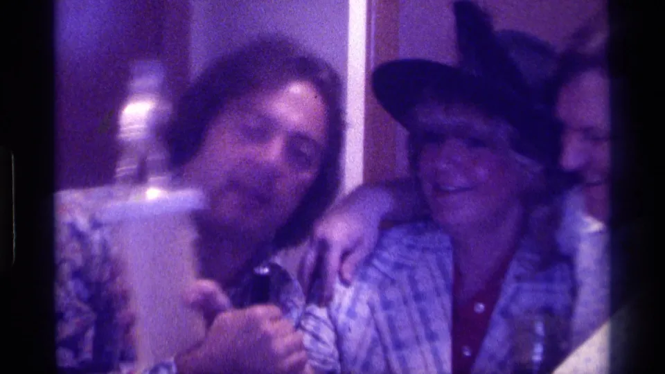 1976 Kentucky 8mm frame of a man holding a trophy near a woman in a black hat, with adults pressed together in a private interior.