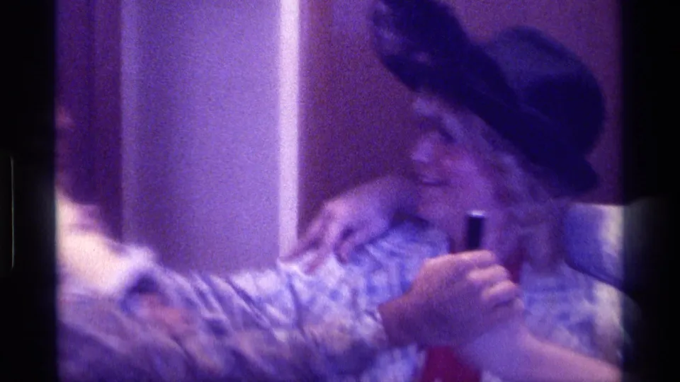 1976 Kentucky home-movie frame showing a woman in a black hat holding a microphone as adults embrace and crowd into the shot indoors.