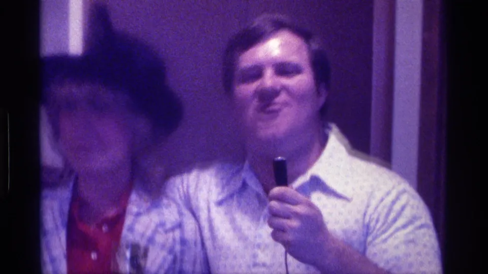 A man with a handheld microphone stands beside a woman in a black hat in an indoor 1976 Kentucky party scene.