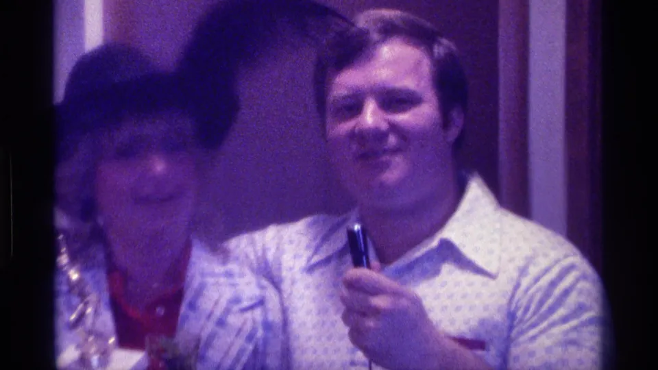 The woman in a black hat appears blurred in the foreground while a man smiles with a microphone in the 1976 Kentucky interior scene.