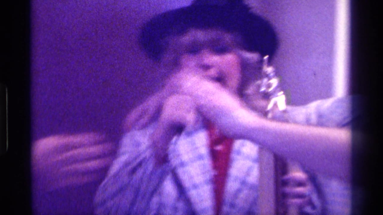 1976 Kentucky home-movie frame of a woman in a feathered hat and plaid jacket holding a trophy and microphone indoors.