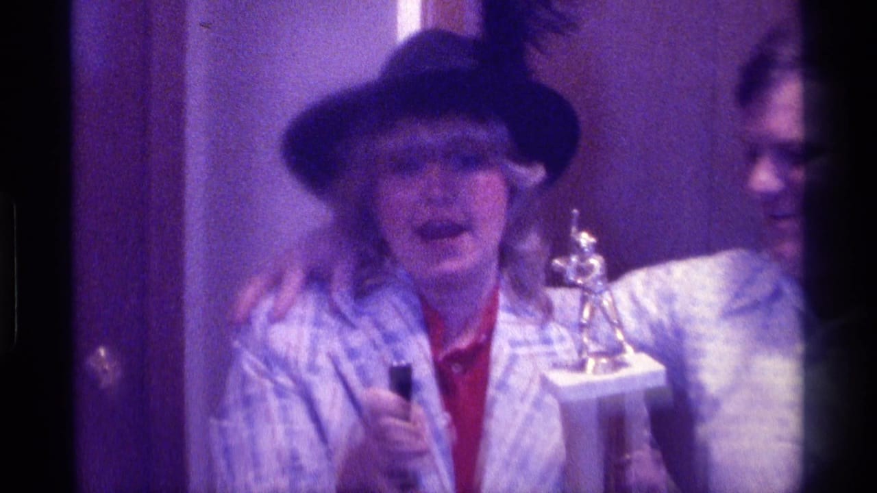 1976 Kentucky home-movie frame showing a woman holding a trophy and microphone beside a smiling man in a patterned shirt.