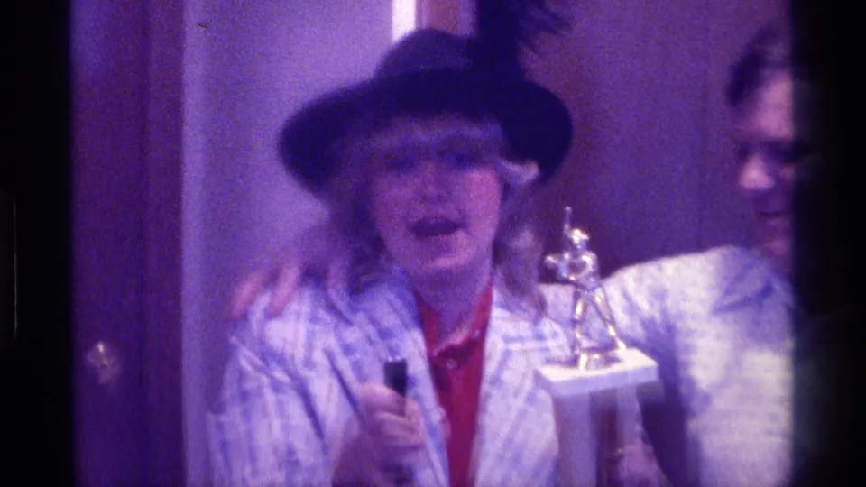 1976 Kentucky home-movie frame showing a woman holding a trophy and microphone beside a smiling man in a patterned shirt.