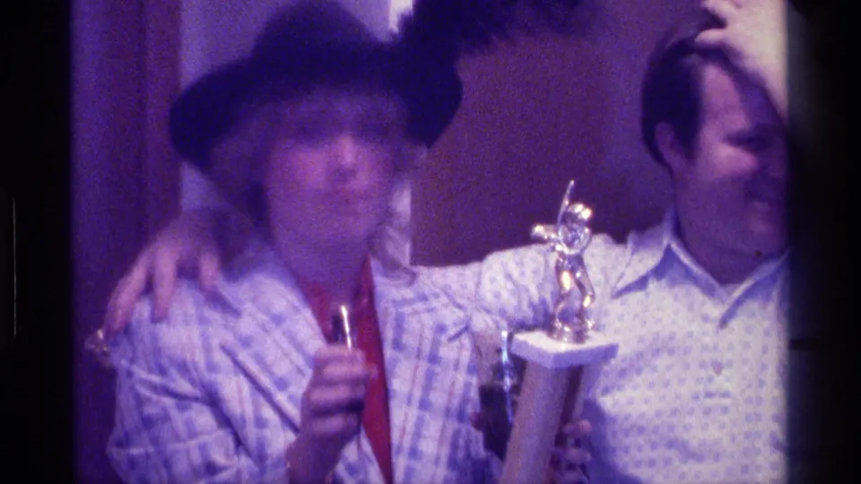 1976 Kentucky indoor home-movie frame of a woman with a trophy and microphone beside a man with his arm around her shoulder.