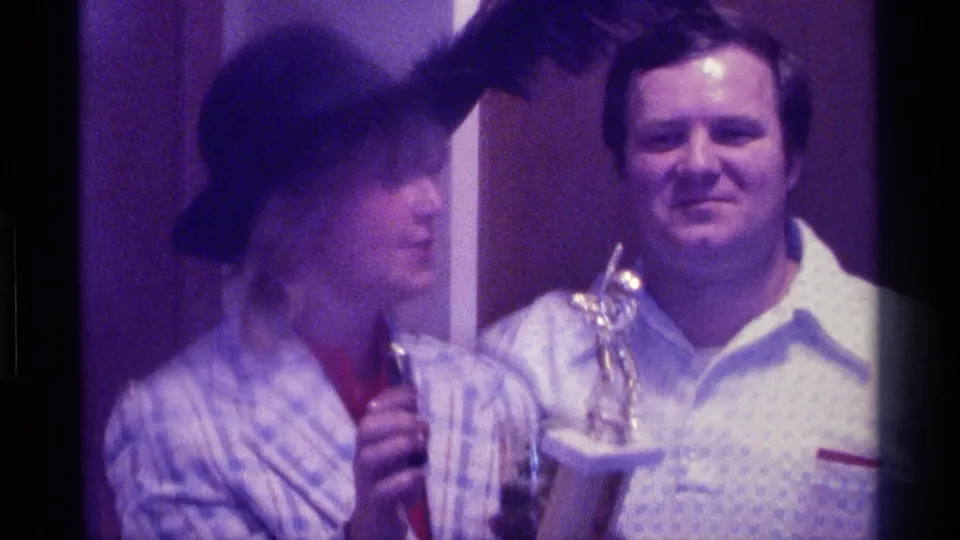 1976 Kentucky home-movie frame with the trophy holder turning toward a smiling man in a dim indoor room.