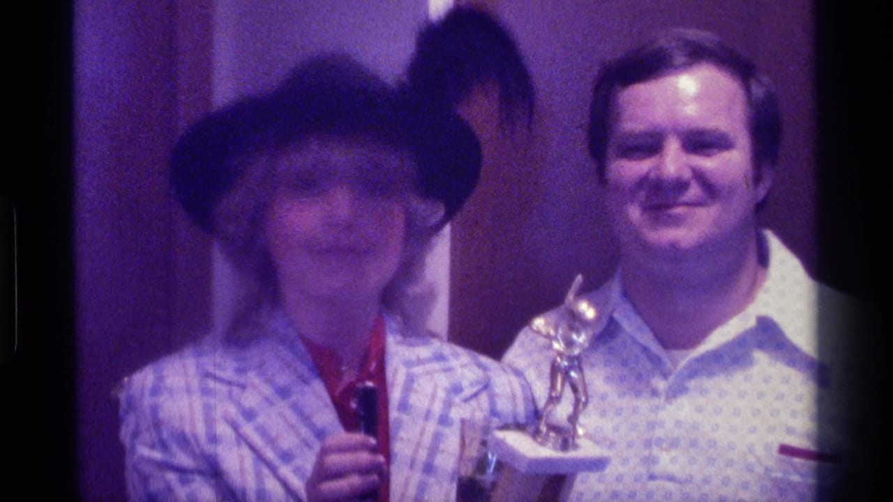 Two adults face the camera as a woman in a black hat holds a microphone beside a man with a trophy.