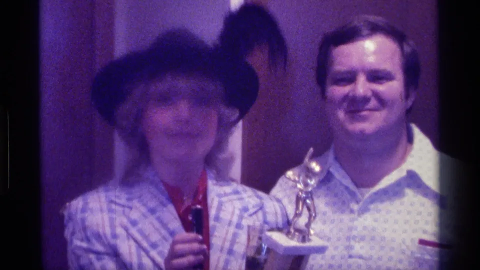 1976 Kentucky home-movie frame showing a woman in a black feathered hat with a microphone beside a man holding a small trophy indoors.
