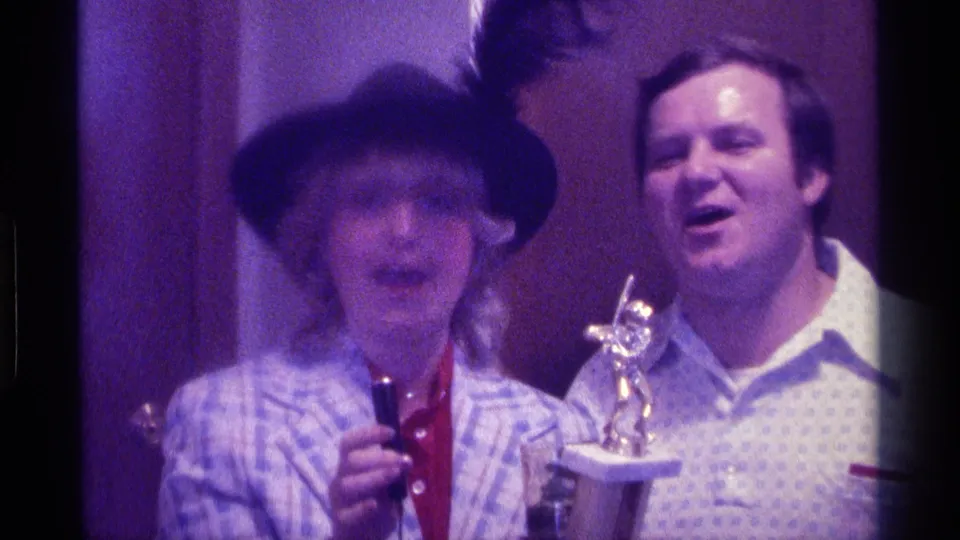 1976 Kentucky frame of a woman speaking with a microphone next to a smiling man holding a trophy in a private interior.