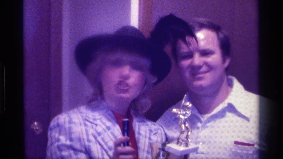 1976 Kentucky home-movie still with the woman leaning forward with a microphone and the man smiling from behind near the trophy.