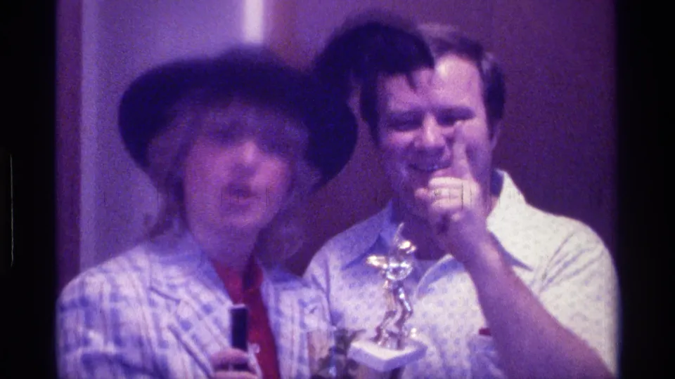 1976 Kentucky frame showing a woman addressing the camera and a man laughing beside a small trophy in an indoor celebration scene.