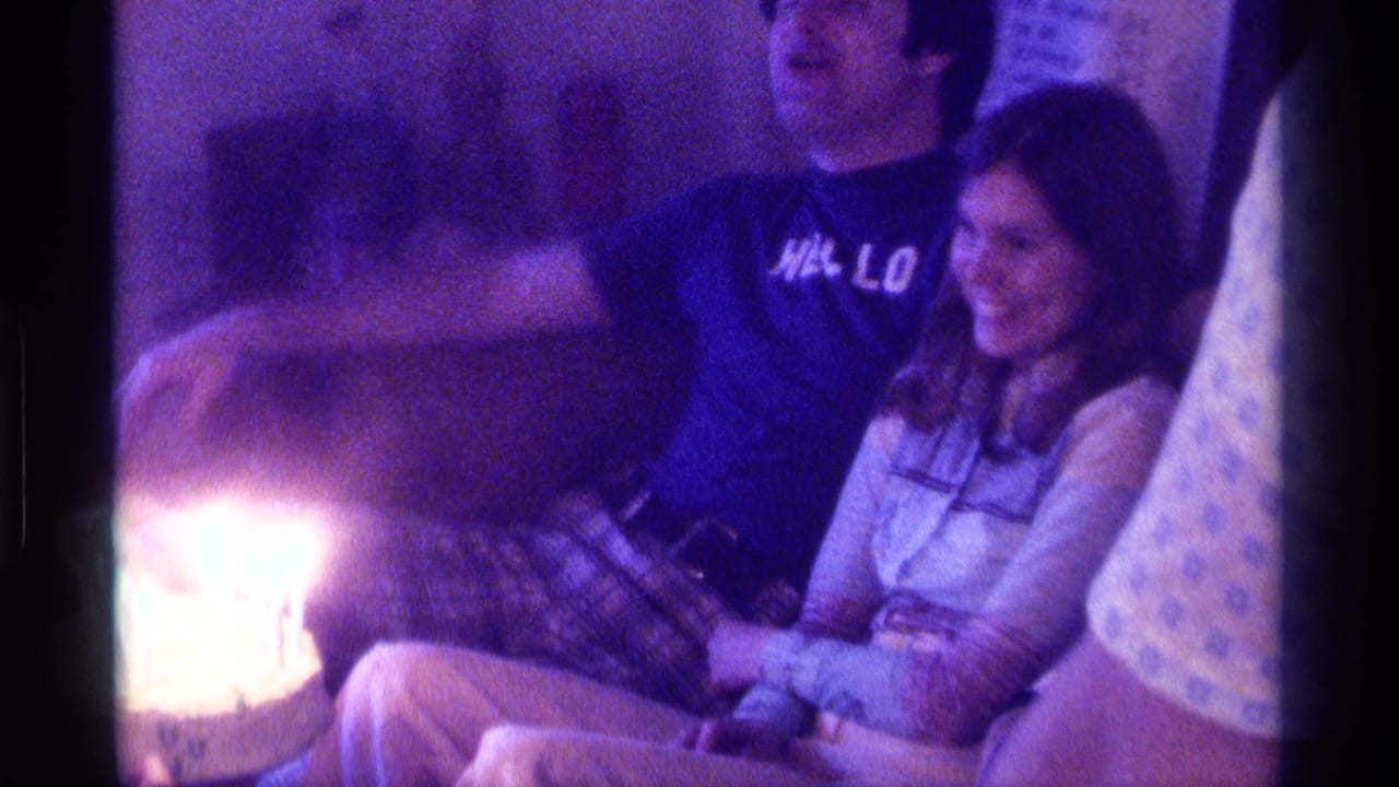 A man and smiling woman sit close together beside lit birthday candles on a couch.