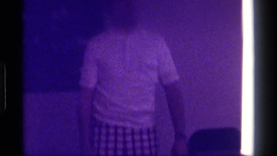 1976 Kentucky 8mm frame showing a person in a white T-shirt and checkered shorts in a purple-tinted living room interior.